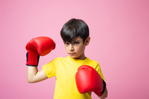 Studio portrait with pink background of a boy with boxing gloves showing off muscle and looking at camera