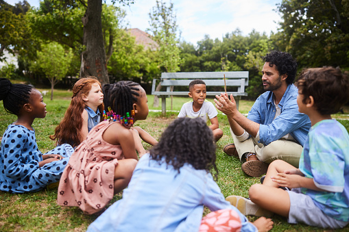 Teacher talking with a diverse group of young students sitting together in a circle on some grass in a park