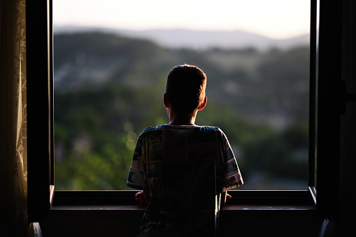 Back of boy looking from window in sunset.
