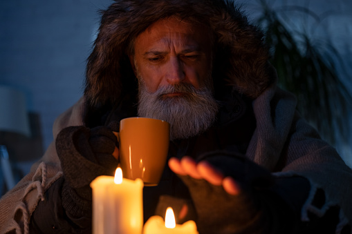 Senior man wearing winter jacket at home with burning candles. High energy prices concept.
