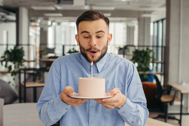 사무실에서 생일을 축하하는 흥분된 남자, 행복해 보이고, 촛불로 케이크를 들고 소원을 빌고 있다. - 253 뉴스 사진 이미지