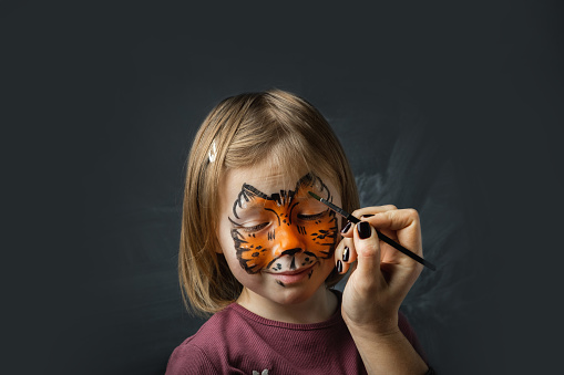 Cute little caucasian girl with tiger face painting on the black background. Close up portrait of little kid with face-painting. Year of a tiger. Happy smiling girl pretending she is a tiger