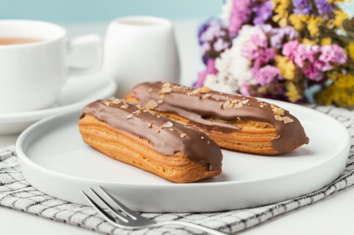 Eclairs with custard and chocolate icing on a white plate. Eclairs with cream filling covered with melted milk chocolate. A cup of tea with traditional French pastry in the morning. French breakfast
