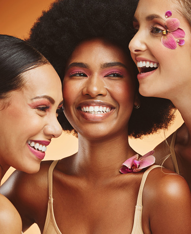 Women, beauty and flowers, makeup and diversity of models on orange studio background. Skincare, face and happy, young and elegant female group posing together with cosmetics, orchids or pink plants.