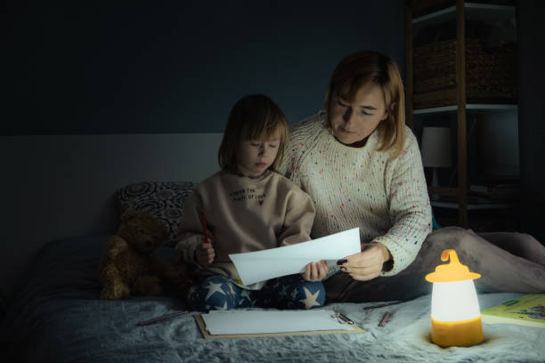 mãe e filha pequena estudando e desenhando em uma escuridão completa durante a falta de eletricidade. menina usa lanterna de acampamento para fazer sua lição de casa durante o apagão. conceito de crise energética - dark little girls child happiness - fotografias e filmes do acervo