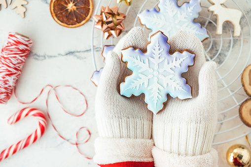 Woman hands wearing white mittens and holding snowflake shaped gingerbread cookie with festive icing on the decorated Christmas background.