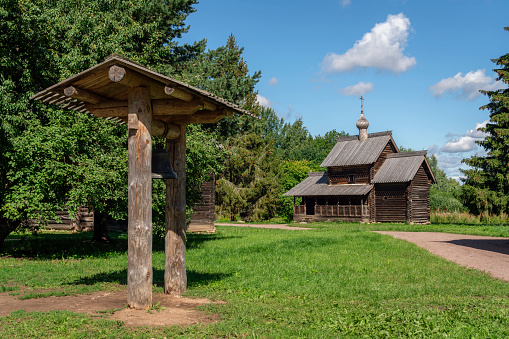 View of the Assumption Church and the wooden belfry with a bell in the foreground at the Novgorod Museum of Folk Wooden Architecture of Vitoslavlitsa on a sunny summer day, Veliky Novgorod, Russia