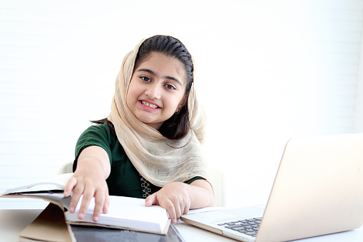 Adorable happy smiling Pakistani Muslim girl with beautiful eyes wearing hijab, studying online, doing homework by using laptop computer, student kid reading book on white background.