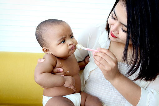 Mother feeding her cute little girl Asian African American baby daughter with spoon of food, infant kid enjoy eating healthy homemade baby food for mom at home.