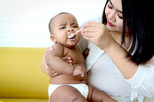 Mother feeding her cute little girl Asian African American baby daughter with spoon of food, infant kid enjoy eating healthy homemade baby food for mom at home.