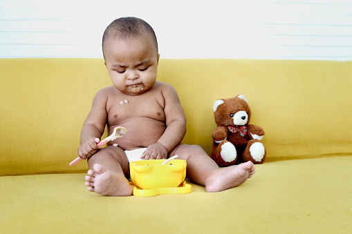 Cute little girl African American baby daughter trying to feed herself with spoon of food, infant kid enjoy eating healthy homemade baby food on yellow sofa at home.
