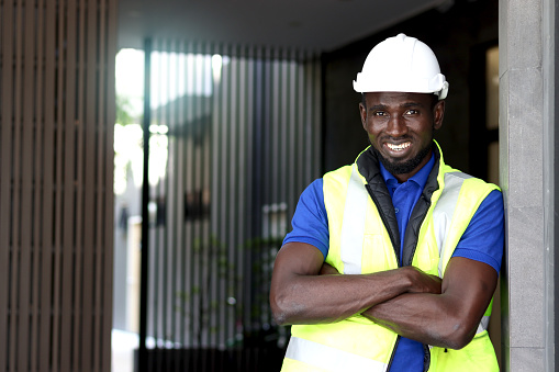 Portrait of young happy smiling African American young engineer worker man wearing safety bright neon color vest and helmet, standing with arms crossed