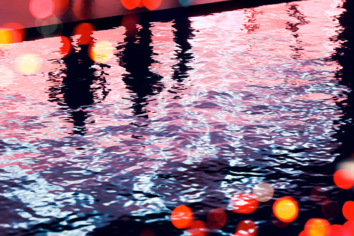 Moody city life: dark silhouettes of three people reflecting on a water surface with waves and ripples, illuminated by neon lights at night, while blurred street lights fill the scene.