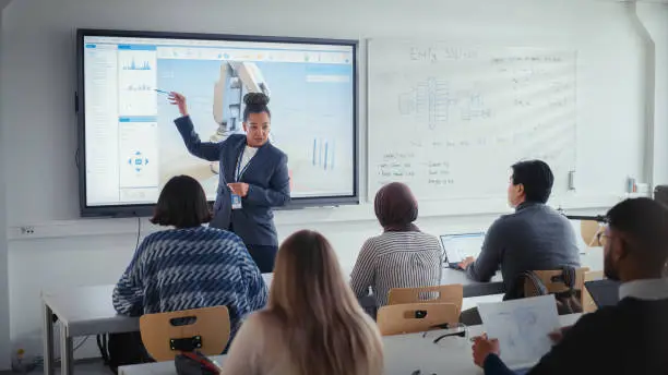 Photo of University Robotics Lecture: Black Teacher Explain Engineering to Students. She Uses Interactive Whiteboard. Diverse Group of Young Engineers Listen in Class About Program Development for Robotic Arm.