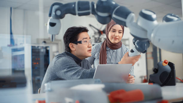 istock Asian Engineer Repositioning and Examining Robotic Arm with Middle Eastern Female Colleague In Robotics Development Laboratory. Research on Design, Operation and Performance of Robotic Mechanism. 1448837374
