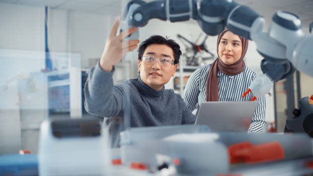 istock Arabic Female Future Engineer Looks At Work of Skilful Developer. Asian Man Explains Core Principles and Examins Robotic Arm. Analysis and Evaluation of Prototypes and Robotic Systems. 1448837396