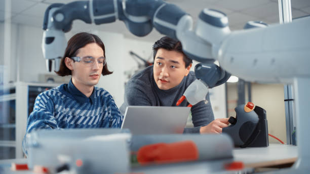 istock Teamwork In Multiethnic Office: Project Manager Supports his Colleague Who is Working on Laptop Computer. Professionals Create Program for Robotic Arm. People Designing Automated Robotic Systems. 1448837419