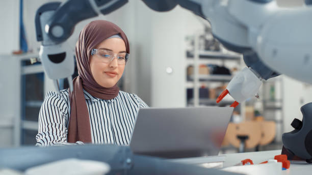 istock Middle Eastern Engineer in Hijab Conducts an In-depth Analysis of the Robot Arm and Programs on Laptop at Modern Office. Optimisation of Autonomous Manufacturing Facility For High Tech Products. 1448837572