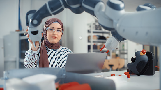 Modern Office: Retrato de una mujer de negocios musulmana que usa hijab y trabaja en un proyecto de ingeniería, codifica en una computadora portátil y cambia la posición de la mano del robot. Ingeniero capacitado con proyecto de inicio. photo