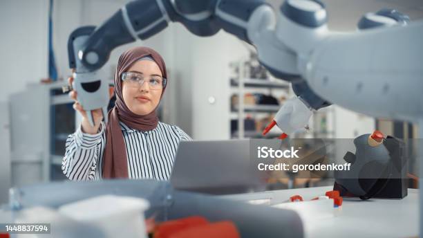 Modern Office Portrait Of Muslim Businesswoman Wearing Hijab And Working On Engineering Project Coding On Laptop And Changing Robot Hand Position Empowered Engineer With Startup Project Stock Photo - Download Image Now