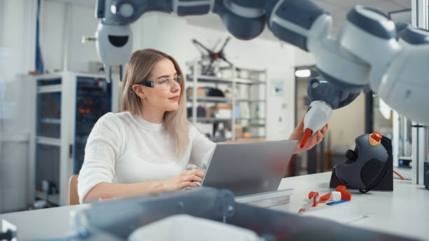 istock Female Automation Engineer Writes Code on Laptop and Moves Bionic Claw At the Factory. Modern and Futuristic Robotic Equipment Starts the New Era in Computer Science, Machine Learning. 1448837618