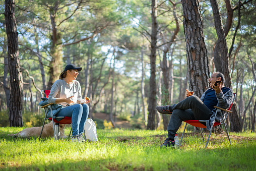Happy middle aged couple relaxing together outdoor sitting on folding chairs at camping table. Man and woman talking, drinking fresh coffee, enjoying summer nature of wild meadow