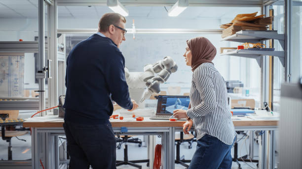 istock Chief Male Engineer Chatting with Arabic Female Computer Science Specialist While Standing Next to the Table with Robotic Hand. Promising Engineering Majors Collaboration Concept. Medium Shot. 1448840006