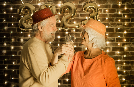 Blurred cheerful senior couple holding champagne flutes having party by night. Happiness lifestyle for mature retired people waiting for new year 2023