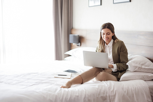 Mid aged brunette white businesswoman sitting on bed in a hotel room using laptop computer during business trip