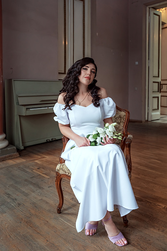 A bride with a bouquet of flowers in a lush white dress sits in a chair and looks into the camera, a portrait of a bride