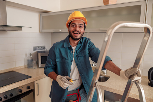 Smiling handyman is standing on ladder in kitchen and holding a screwdriver. High quality photo