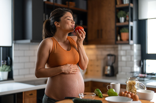 Young attractive pregnant woman smelling tomato while preparing a delicious meal in the kitchen.