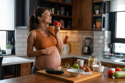 Young pregnant woman holding and smelling a fresh tomato with the eyes closed in the kitchen at home. Touching her belly, enjoying pregnancy, making breakfast.