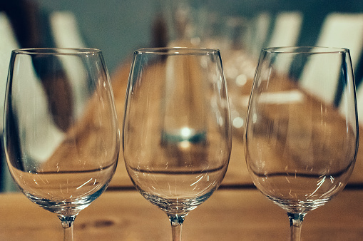 Close-up of three empty glasses on a wooden table, served for wine tasting event. White chairs in the blurred background. Bar or restaurant interior, subdued light. Selective focus, film grain effect