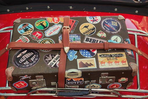An old suitcase with country tags tied to the trunk of an old red car. Old vintage suitcase decorated with stickers with flags of different countries  Gökçeada Imbros island Çanakkale, Turkey June 16, 2021