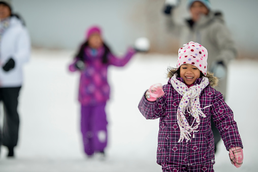 A small Asian family of four play together in the snow on a cold winters day.  They are each dressed in warm snow suits as they run around and throw snowballs at one another.
