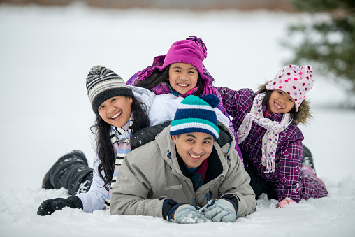 A small Asian family of four stack on top of one another in the snow as they pose for a portrait on a cold winters day.  They are each dressed warmly in snowsuits and smiling as they enjoy each others company and the fresh air.