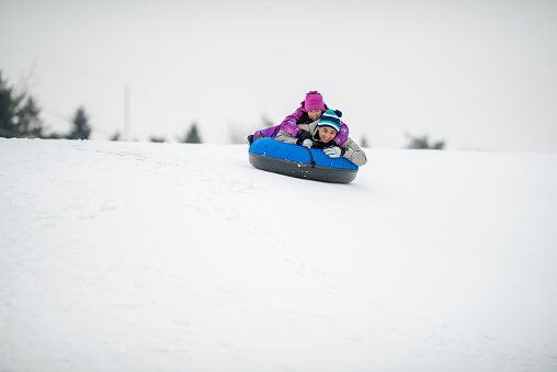 A young Asian girl lays on her Dad's back on a snow tube as they ride down the hill together.  They are each dressed warmly in snowsuits as they enjoy each others company and the fresh air.