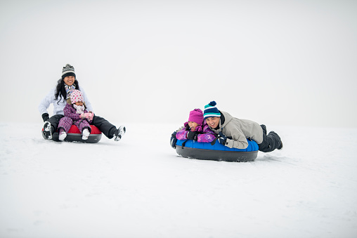 A small Asian family of four spend some time outside together tubing down a hill on a winters day.  They are each dressed warmly in snow suits as they laugh and enjoy each others company.