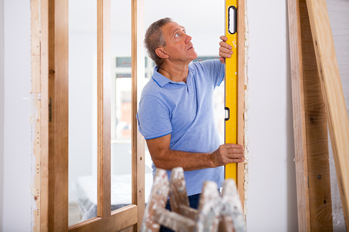 Focused middle-aged man using a waterpass on a doorjamb during installation of a door at home