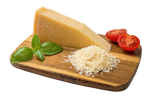 Whole and grated parmesan on a cutting board cutout. Grana padano wedge, grated cheese, basil and halved tomato on a wooden shopping board isolated on a white background. Dairy product. Front view.