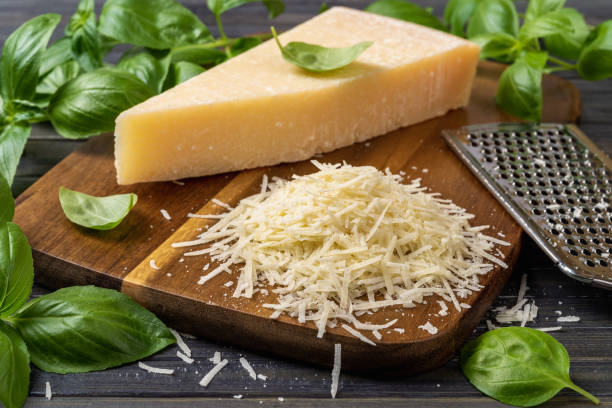 Shredded parmesan cheese and grater on a cutting board. Grana padano cheese whole wedge and grated, stainless steel grater and green basil herb over wooden background. Hard cheese. stock photo