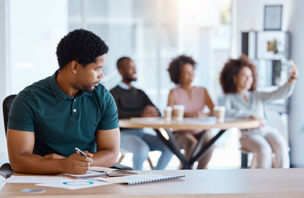 istock Lonely, alone and African businessman in the office working on a project while colleagues take selfie. Reject, sad and antisocial professional employee sitting at his desk as outcast in the workplace 1448898808