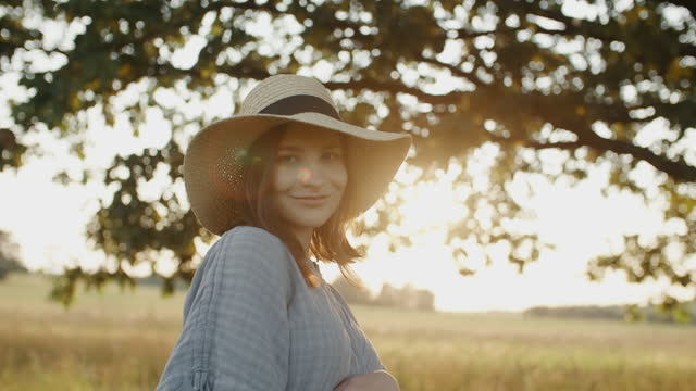 Pregnant woman wearing linen dress and straw hat standing by oak tree in meadow in sunset light
