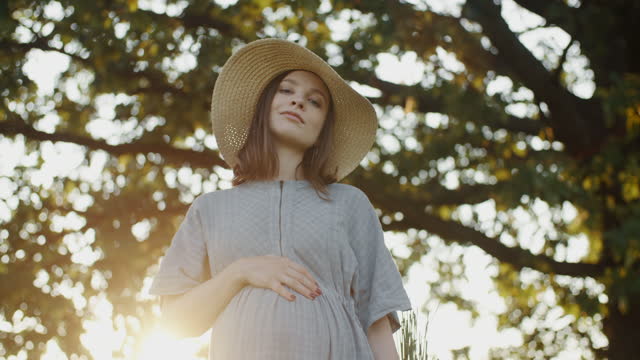 Happy pregnant woman wearing linen dress and hat standing by oak tree in meadow during sunset