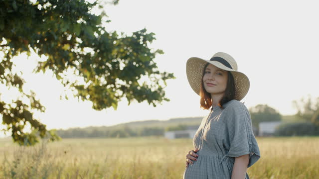 Pregnant woman in linen dress and hat posing near oak tree in meadow outdoors during sunset