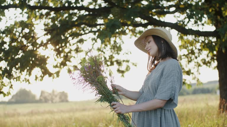 Pregnant woman standing outdoors with flower bouquet by oak tree in meadow during sunset