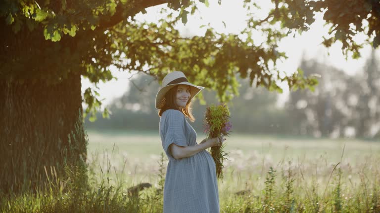 Pregnant woman in linen dress standing with flowers bouquet outdoors by oak tree in the meadow during sunset