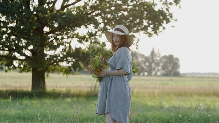 Pregnant woman in linen dress and hat walks with wild flowers bouquet outdoors by oak tree in meadow on sunset