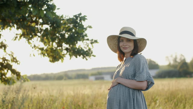 Pregnant woman in linen dress and hat posing near oak tree in meadow outdoors during sunset